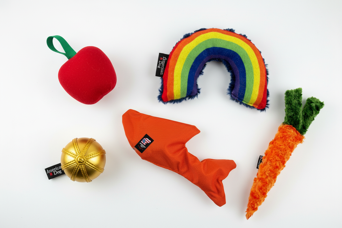 Colorful collection of Lucky Dog $7 USA-made dog toys including apple tug, rainbow plushie, golden snitch tug, fish bungee tug, and carrot plushie arranged on a white surface